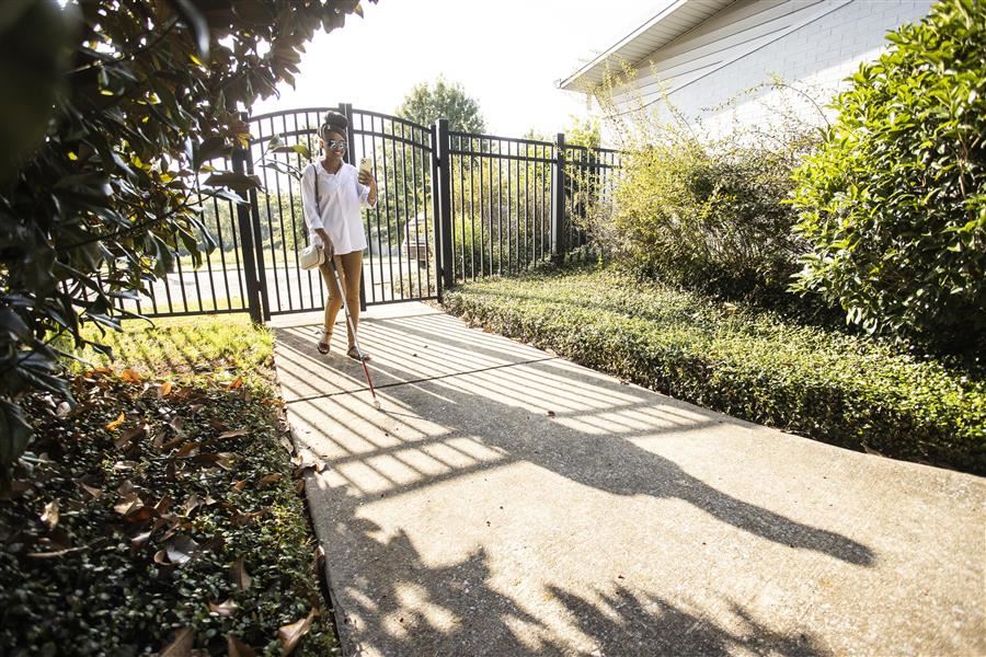   Jonesti Steele walks down a sidewalk between buildings using a phone and white cane to navigate. 