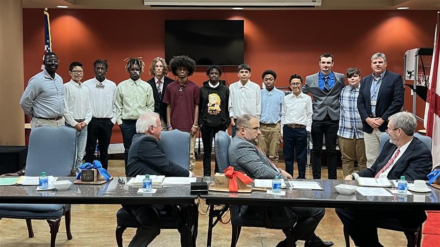   The Boy's Track Team and Coaches smile to camera behind three trustees, seated, looking on.