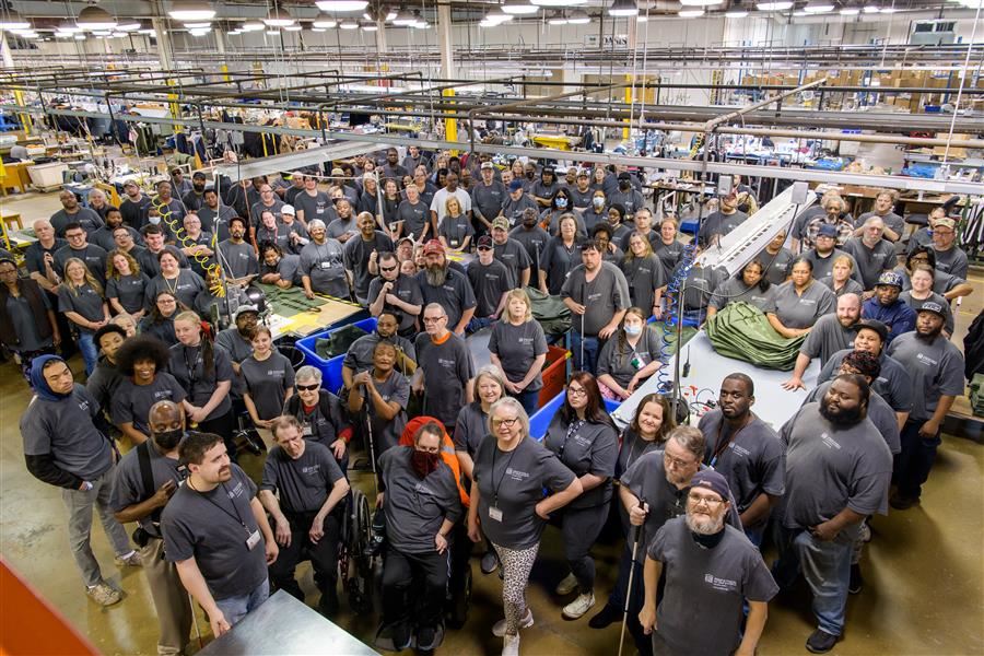 All staff members from Alabama Industries for the Blind stand on the production room floor and smile up to camera. 