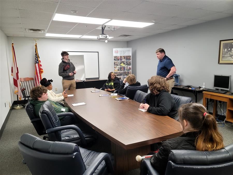 Students sit around a table; One student stands at the head of the table; They are looking at A.I.B. Executive Director 