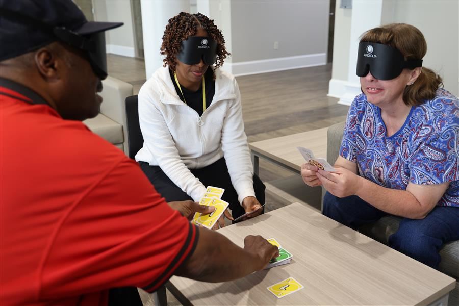  Three adults wearing mindfold blind folds are seated around a table while playing Uno.
