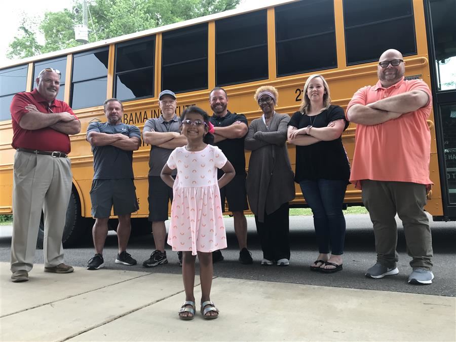 A I D B transportation workers stand in a group behind an A S B student. In the background is an A I D B school bus.
