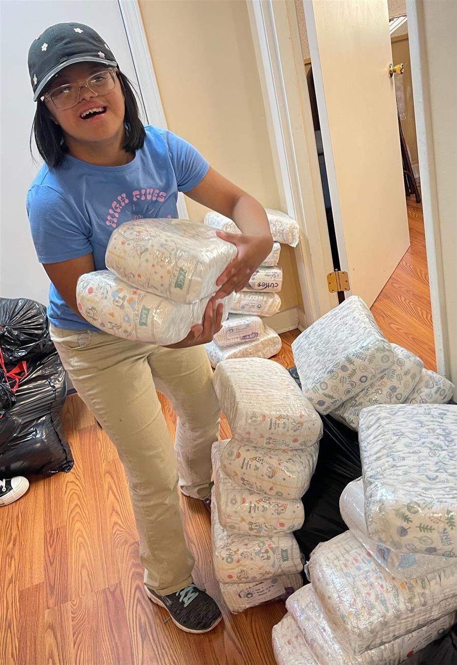 A student holds two sets of diapers as she organizes them at Diaper Row. She smiles to camera. 