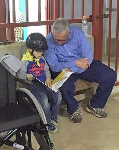 A male volunteer helps a young boy waiting to ride a horse at the MGHSE arena read a book A male volunteer helps a young boy waiting to ride a horse at the MGHSE arena read a book