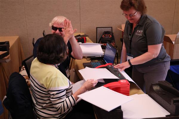 CATT trainer Susie Thomas assisting two Deaf Blind Limitless conference attendees load braille paper into a page blaster before embossing