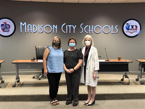 Three women pose for a picture in a room containing a sign for Madison County Schools.