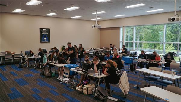 In a large classroom, students are working on computer assignments and receiving help from mentors and educators