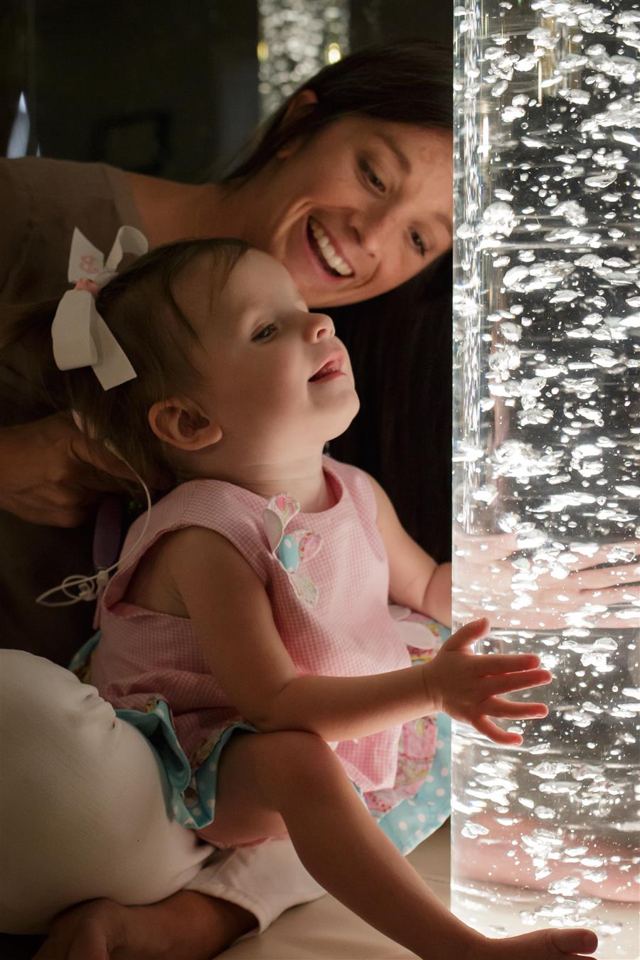 A toddler looks up at bubbles while seated in her mothers lap.